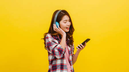Young Asian lady wearing wireless headphones listening to music from smartphone with cheerful expression in casual clothing and looking at camera over yellow background. Facial expression concept.の写真素材