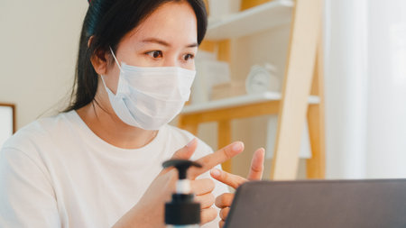 Asian business woman wearing mask using laptop working call video conference with customer in living...