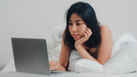 Young Asian woman using laptop checking social media feeling happy smiling while lying on bed after...
