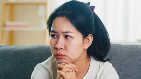 Depressed crying Asian business woman stressed with headache sitting on sofa in living room at house .の写真素材