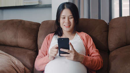 Young Asian Pregnant woman using mobile phone search pregnancy information. Mom feeling happy smiling positive and peaceful while take care her child lying on sofa in living room at home concept.の写真素材