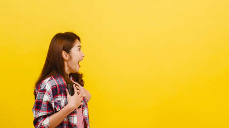 Portrait of surprised Asian female using mobile phone with positive expression, dressed in casual clothing and looking at camera over yellow background. Happy adorable glad woman rejoices success.の写真素材