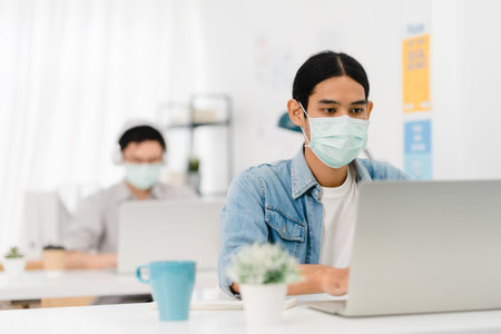 Asia businessman entrepreneur wearing medical face mask for social distancing in new normal situation for virus prevention while using laptop back at work in office. Lifestyle after corona virus.の写真素材