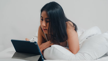 Young Asian woman using tablet checking social media feeling happy smiling while lying on bed after...