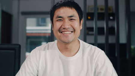 Portrait Asian Chinese man feeling happy funny at home. Young male relax toothy smile looking to camera sitting on table in living room at house in the morning concept.の写真素材