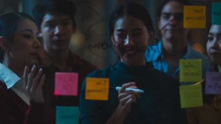 Asia young creative people discussing business brainstorming meeting working together sharing data and writing glass wall with sticky notes feeling happy in night office. Coworker teamwork concept.の写真素材