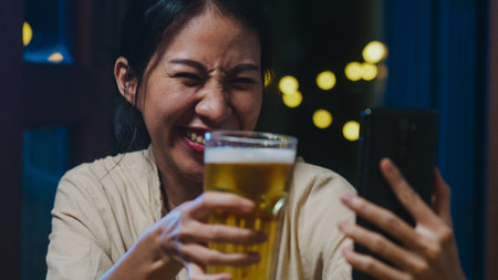Young Asia lady drinking beer having fun happy moment night party New Year event online celebration via video call by phone at home at night. Social distancing, quarantine for coronavirus prevention.の写真素材
