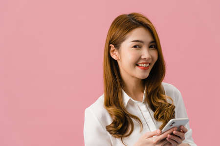 Surprised young Asia lady using mobile phone with positive expression, smiles broadly, dressed in casual clothing and looking at camera on pink background. Happy adorable glad woman rejoices success.の写真素材