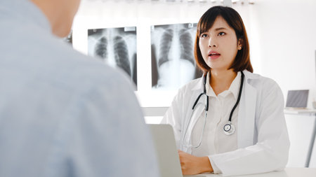 Young Asia female doctor in white medical uniform using computer laptop is delivering great news talk discuss results or symptoms with male patient sit at desk in health clinic or hospital office.の写真素材