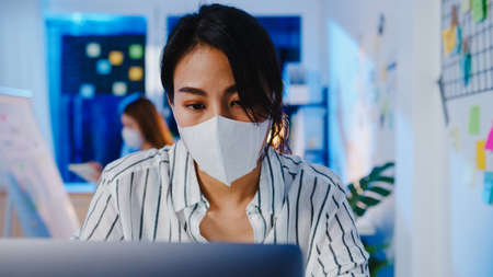 Happy Asia businesswoman wearing medical face mask for social distancing in new normal situation for virus prevention while using laptop back at work in office night.の写真素材