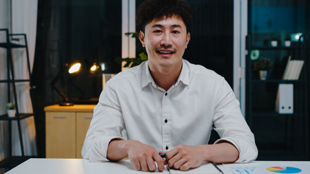 Asia businessman social distancing in new normal for virus prevention looking at camera presentation to colleague about plan in video call while work in office night.の写真素材