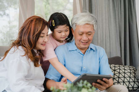 Asian grandparents and granddaughter using tablet at home. Senior Chinese, grandpa and grandma happy spend family time relax with young girl checking social media, lying on sofa in living room conceptの写真素材