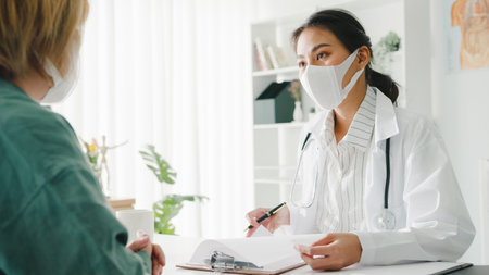Young Asia lady doctor wear protective mask using clipboard is delivering great news talk discuss results or symptoms with girl patient in hospital office.の写真素材