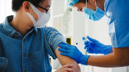 Young Asia lady nurse giving  flu antivirus vaccine shot to senior male patient wear face mask protection from virus disease at health clinic or hospital office. Vaccination concept.の写真素材
