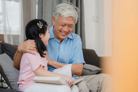 Asian grandfather relax at home. Senior Chinese, grandpa happy relax with young granddaughter girl enjoy read books and do homework together in living room concept.の写真素材