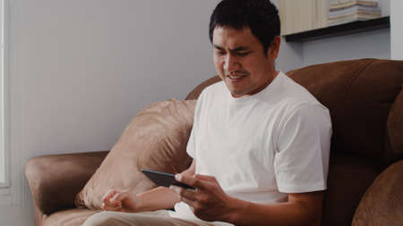 Young Asian man using mobile phone playing video games in television in living room, male feeling happy using relax time lying on sofa at home. Men play games relax at home concept.の写真素材