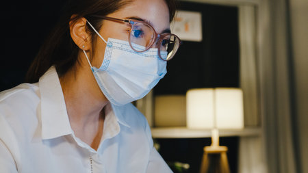 Freelance Asia lady wear medical face mask use laptop hard work in living room at house. Working from home overload at night, remotely work, social distancing, quarantine for  virus prevention.の写真素材