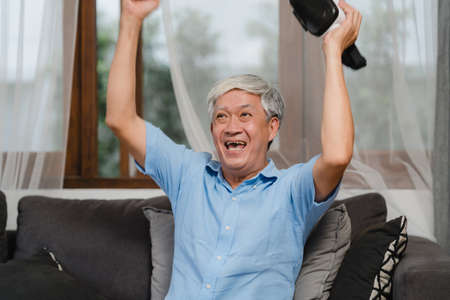 Asian senior men play games at home. Asian senior older Chinese male happy fun and virtual reality, VR playing games while lying sofa in living room at home concept.の写真素材