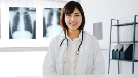 Confident young Asia female doctor in white medical uniform with stethoscope looking at camera and smiling while video conference call with patient in health hospital. Consulting and therapy concept.の写真素材