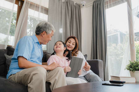 Asian grandparents and granddaughter using tablet at home. Senior Chinese, grandpa and grandma happy spend family time relax with young girl checking social media, lying on sofa in living room conceptの写真素材