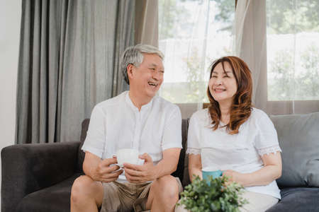 Asian senior couple relax at home. Asian Senior Chinese grandparents, husband and wife smile talking and drinking coffee while lying on sofa in living room at home concept.の写真素材
