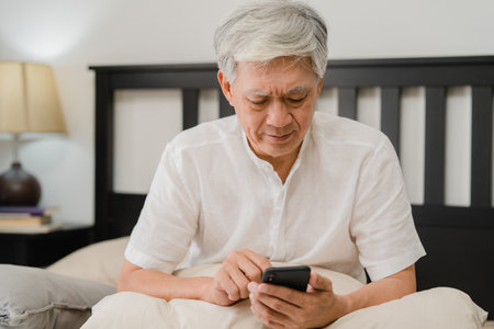 Asian senior men using mobile phone at home. Asian Senior Chinese male search information about how to good health on internet while lying on bed in bedroom at home in the morning concept.の写真素材