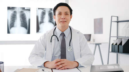 Confident young Asia male doctor in white medical uniform with stethoscope looking at camera and smiling while video conference call with patient in health hospital. Consulting and therapy concept.の写真素材