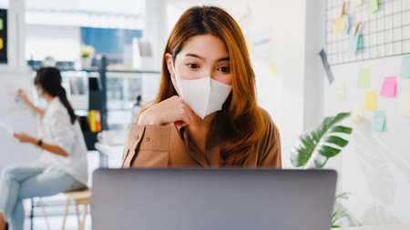 Asia businesswoman wear face mask for social distancing in new normal situation for virus prevention while using laptop presentation to colleagues about plan in video call while work in office.の写真素材