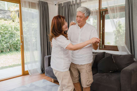 Asian elderly couple dancing together while listen to music in living room at home, sweet couple enjoy love moment while having fun when relaxed at home. Lifestyle senior family relax at home concept.の写真素材