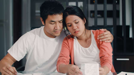 Young Asian Pregnant couple records of income and expenses at home. Mom worried, serious, stress while record budget, tax, financial document working in living room at home concept.の写真素材