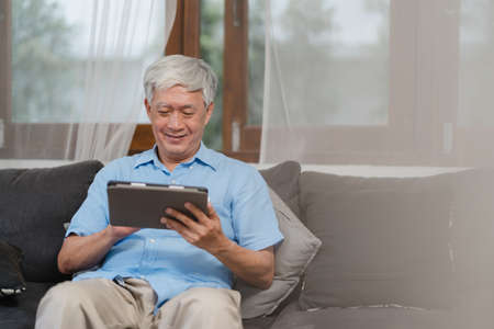 Asian senior men using tablet at home. Asian Senior Chinese male search information about how to good health on internet while lying on sofa in living room at home concept.の写真素材