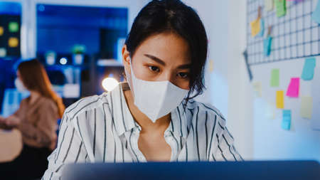 Happy Asia businesswoman wearing medical face mask for social distancing in new normal situation for virus prevention while using laptop back at work in office nightの写真素材