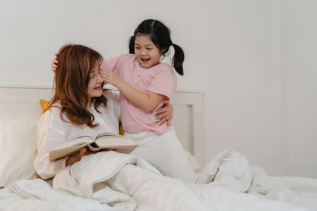 Asian grandmother relax at home. Senior Chinese, grandma happy relax with young granddaughter girl enjoy close her eyes surprise playing together lying on bed in bedroom at home at night concept.の写真素材