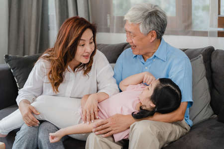 Asian grandparents talking with granddaughter at home. Senior Chinese, old generation, grandfather and grandmother using family time relax with young girl kid lying on sofa in living room concept.の写真素材