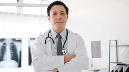 Young Asia male doctor in white medical uniform with stethoscope looking at camera, smile and arms crossed while video conference call with patient in health hospital. Consulting and therapy concept.の写真素材