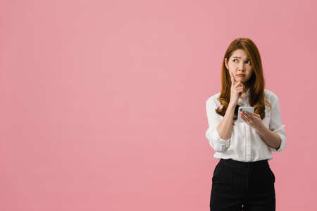 Thinking dreaming young Asia lady using phone with positive expression, dressed in casual clothing feeling happiness and stand isolated on pink background. Happy adorable glad woman rejoices success.の写真素材