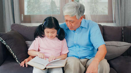 Asian grandfather relax at home. Senior Chinese, grandpa happy relax with young granddaughter girl enjoy read books and do homework together in living room concept.の写真素材