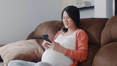 Young Asian Pregnant woman using mobile phone search pregnancy information. Mom feeling happy smiling positive and peaceful while take care her child lying on sofa in living room at home concept.の写真素材