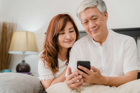 Asian senior couple using mobile phone at home. Asian Senior Chinese grandparents, husband and wife happy after wake up, watching movie lying on bed in bedroom at home in the morning concept.の写真素材