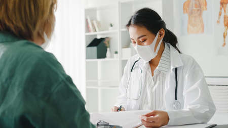 Young Asia lady doctor wear protective mask using clipboard is delivering great news talk discuss results or symptoms with girl patient in hospital office.の写真素材