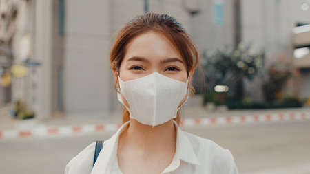 Successful young Asia businesswoman in fashion office clothes wear medical face mask smiling and looking at camera while happy standing alone outdoors in urban modern city. Business on the go concept.の写真素材