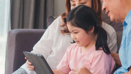 Asian grandparents and granddaughter using tablet at home. Senior Chinese, grandpa and grandma happy spend family time relax with young girl checking social media, lying on sofa in living room conceptの写真素材