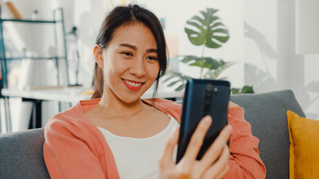Beautiful asian lady smiling and sitting on sofa call phone talk say hi with family at home. Stay at...