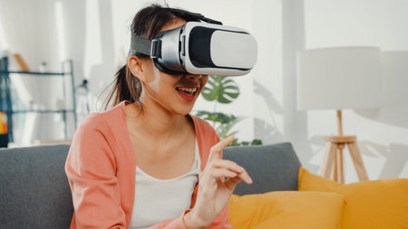 Asian lady wearing headset glasses of virtual reality gesticulating hand sitting on couch in living...