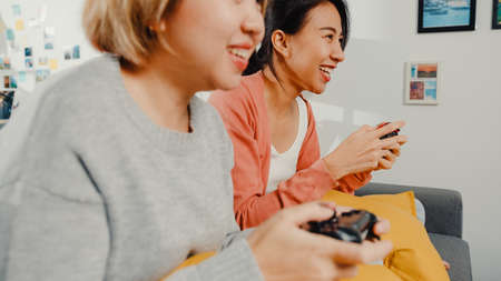 Lesbian LGBTQ women couple play video game at home. Young Asia lady using wireless controller having funny happy moment together on sofa in living room. They have great and fun time celebrate holiday.の写真素材
