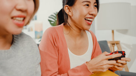 Lesbian LGBTQ women couple play video game at home. Young Asia lady using wireless controller having...