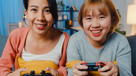 Lesbian LGBTQ women couple play video game at home. Young Asia lady using wireless controller having...