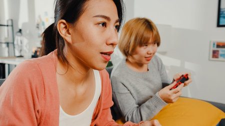 Lesbian LGBTQ women couple play video game at home. Young Asia lady using wireless controller having...