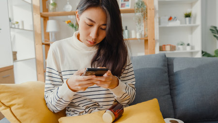 Sick young Asian woman hold medicine sit on couch take photo sent to doctor at home. Girl take medicine after doctor order, quarantine at houseの写真素材