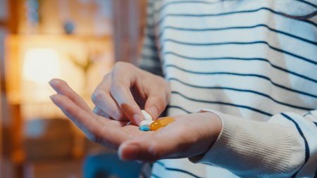 Sick asian young lady holding pill take medicine sit on couch in living room at home night. Girl...